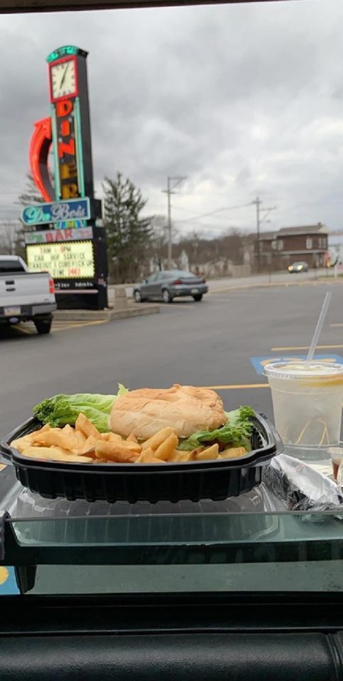 A takeout meal with a sandwich, fries, and a drink, with a diner sign and cloudy sky in the background.
