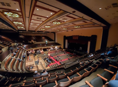 A spacious theater interior with empty seats, a stage, and a few audience members in the background.