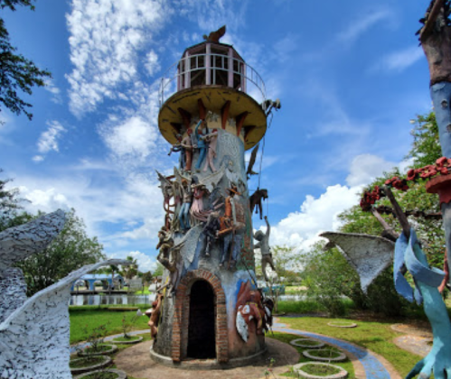 Colorful lighthouse sculpture surrounded by greenery and blue skies, featuring intricate figures and artistic designs.