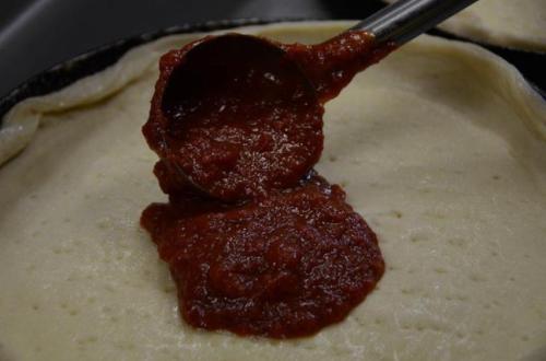 A ladle scoops marinara sauce onto a raw pizza dough in a pan.