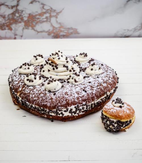 A large chocolate cake topped with whipped cream and chocolate sprinkles, alongside a smaller filled donut.