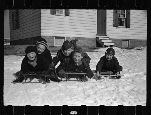 Five children in winter clothing sledding on snow, smiling and lying on sleds in front of a house.