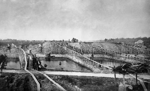 Historic black and white photo of a large excavation site with wooden structures and water-filled trenches.
