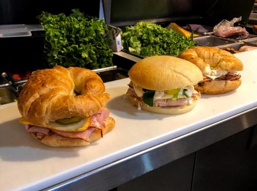 Three sandwiches on a counter: a croissant with ham and cheese, a sandwich with turkey and toppings, and a soft bun sandwich.