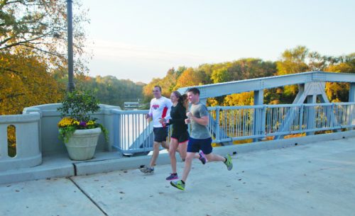 Three runners jog across a bridge with autumn trees in the background, enjoying a sunny morning.