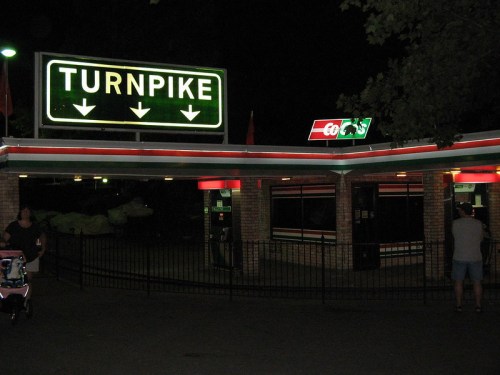 A brightly lit turnpike entrance at night, featuring a sign and a small building with red and white accents.