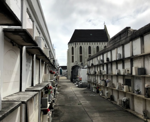 A narrow path between mausoleums in a cemetery, with a church visible in the background under a cloudy sky.
