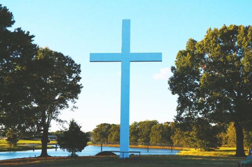 A large white cross stands in a grassy area near a calm pond, surrounded by trees under a clear blue sky.