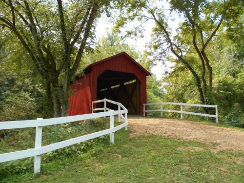 A red covered bridge surrounded by trees and a white fence on a grassy path.