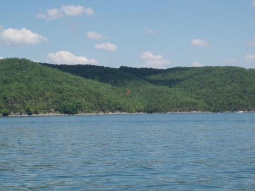 A serene view of a green hillside reflected in calm water under a clear blue sky with a few clouds.