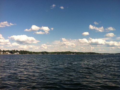 A serene lake scene with gentle waves, under a blue sky filled with fluffy white clouds and a distant tree-lined shore.