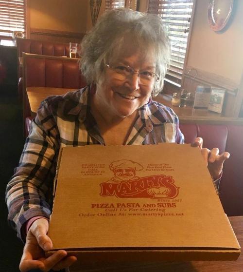 A smiling woman holds a large pizza box from Marty's Pizza Pasta and Subs in a cozy restaurant setting.