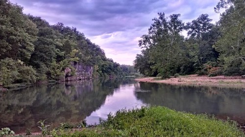 A serene river scene surrounded by lush greenery and rocky cliffs under a cloudy sky. Reflections on the water.