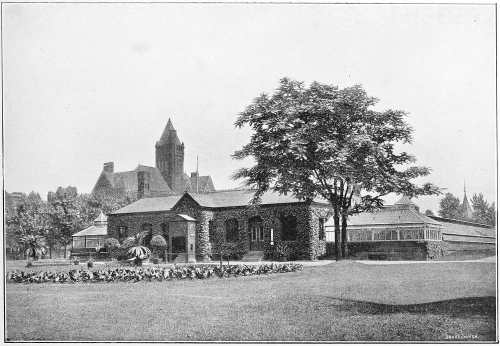 Historic building with a large tree and garden, featuring a stone structure and a tower in the background.