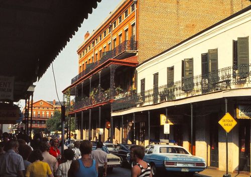 A busy street scene with people walking, vintage cars, and historic buildings featuring balconies in a lively urban setting.