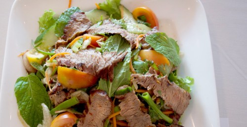 A fresh salad with sliced beef, mixed greens, tomatoes, cucumbers, and herbs in a white bowl.
