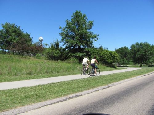 Two people ride bicycles on a paved path surrounded by greenery under a clear blue sky.