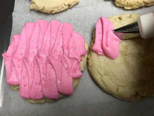A close-up of two cookies, one being decorated with pink frosting using a piping bag.