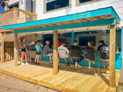 A vibrant outdoor bar with patrons seated at the counter, enjoying drinks under a blue awning on a sunny day.
