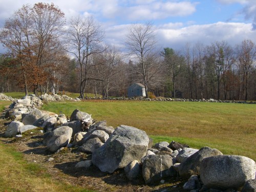 A grassy field with a stone wall, bare trees, and a small blue house in the background under a partly cloudy sky.
