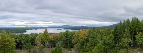 A panoramic view of a lake surrounded by lush green trees and distant mountains under a cloudy sky.