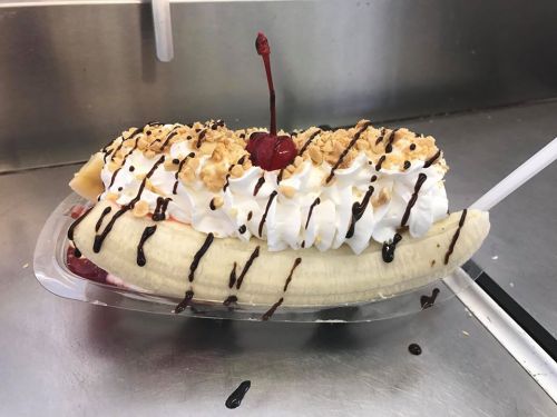 A banana split topped with whipped cream, chocolate drizzle, crushed nuts, and a cherry on top.