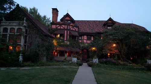 A large, ivy-covered mansion with warm lights glowing from the windows, set in a lush garden at dusk.