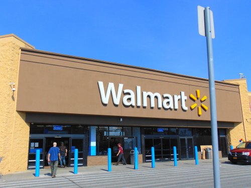 Exterior of a Walmart store with blue poles in front and clear blue sky above. Shoppers are entering and exiting.