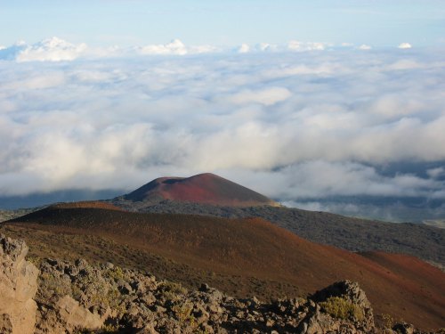 A volcanic landscape with a reddish peak rising above a sea of clouds under a blue sky. Rocky terrain in the foreground.