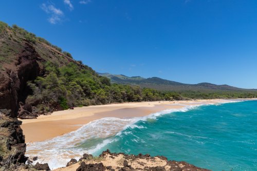 A scenic beach with golden sand, turquoise waves, and lush green hills under a clear blue sky.