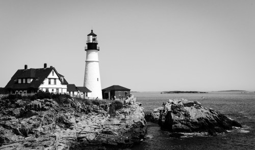 A black and white image of a lighthouse beside a rocky shore, with a house nearby and calm waters in the background.