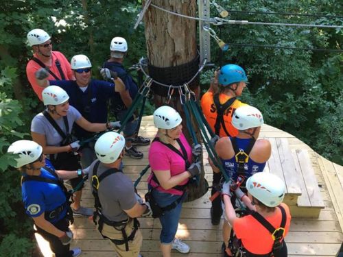 A group of people in helmets and harnesses preparing for a zipline activity on a wooden platform in a forested area.