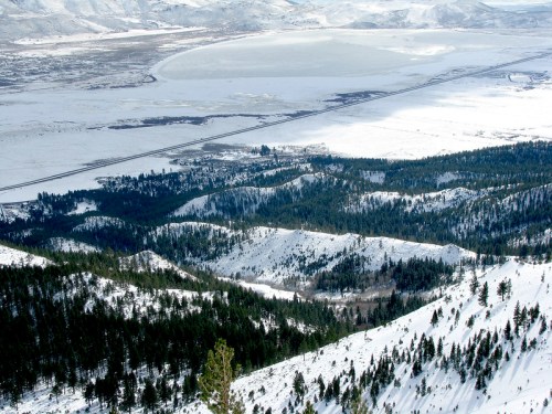 A snowy landscape with rolling hills, pine trees, and a large lake in the distance under a clear sky.