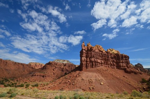 A rugged red rock formation rises against a blue sky with scattered clouds in a desert landscape.