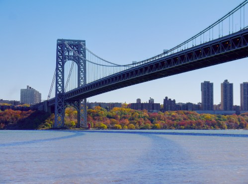 The George Washington Bridge spans a river, with colorful autumn trees and city buildings in the background.