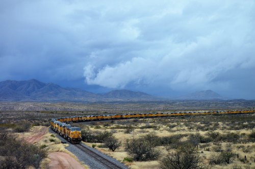 A long freight train curves through a desert landscape under a cloudy sky, with mountains in the background.