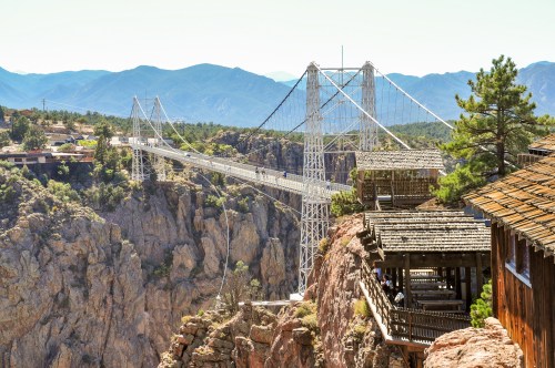 A suspension bridge spans a canyon, surrounded by mountains and trees, with a wooden structure nearby.