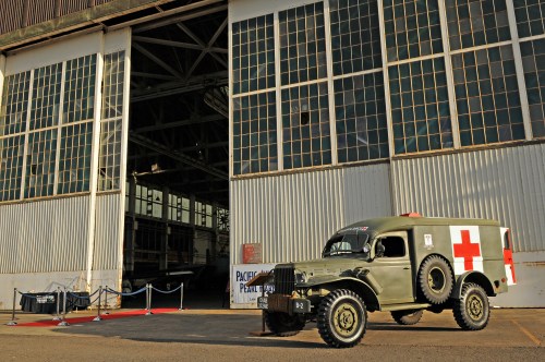 A vintage military ambulance parked outside a large hangar with a red cross symbol, ready for an event.