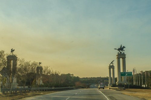 Entrance to a road with tall columns featuring horse statues, leading to Fort Bragg, under a golden sky.