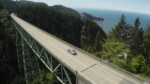 A car drives along a scenic bridge surrounded by lush trees, overlooking a coastal view.