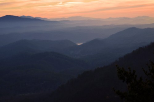 A serene landscape of rolling mountains at sunset, with layers of mist and soft colors in the sky.