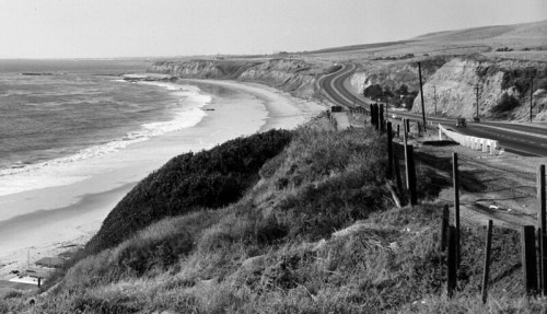 Black and white coastal landscape featuring a winding road along the shore and cliffs overlooking the ocean.