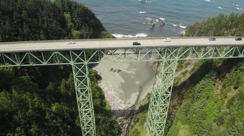 Aerial view of a green bridge spanning a rocky coastline with cars driving above and lush trees surrounding the area.