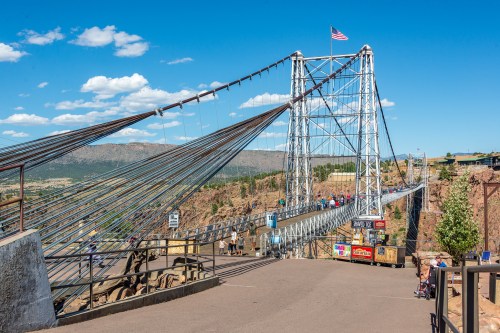A suspension bridge with cables, surrounded by mountains and blue sky, with visitors walking across.