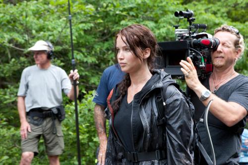 A film crew on set in a forest, with a focused actress in a black jacket and crew members operating equipment.