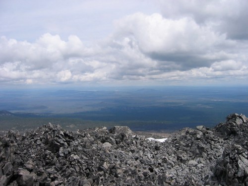 A rocky mountain summit with a panoramic view of distant hills and a cloudy sky.