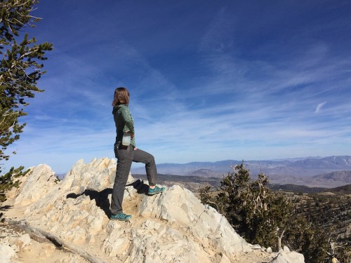 A person stands on a rocky outcrop, gazing at a vast mountain landscape under a clear blue sky.
