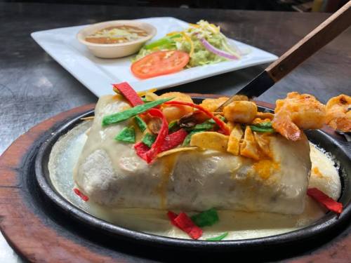 A large burrito topped with colorful toppings, served on a sizzling plate with a side of salad and beans.
