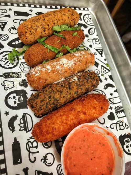 A tray with five different croquettes garnished with herbs, accompanied by a small cup of red dipping sauce.