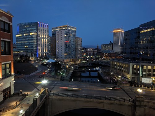 City skyline at dusk, featuring modern buildings, a river, and light trails from moving vehicles on a bridge.